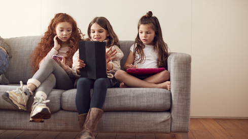 Tre barn sitter på en sofa og bruker digitale enheter. Det ene barnet til venstre holder en smarttelefon, barnet i midten leser på en nettbrett, og barnet til høyre holder et annet nettbrett. De sitter komfortabelt i et hjemmemiljø med en nøytral farget bakgrunn.